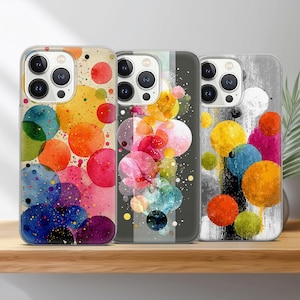 May include: Three phone cases with colorful abstract designs. The cases feature vibrant watercolor-style circles in shades of yellow, orange, pink, blue, and purple. Each case has a different background, including a neutral beige, a dark gray, and a light gray.