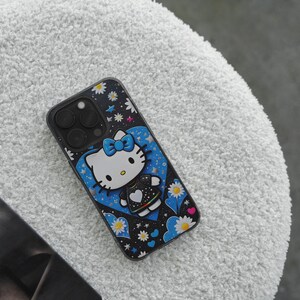 May include: Black phone case with a Hello Kitty design. The case has a black background with white daisies and stars. Hello Kitty is in a blue heart with a blue bow. The case is on a white, textured surface.