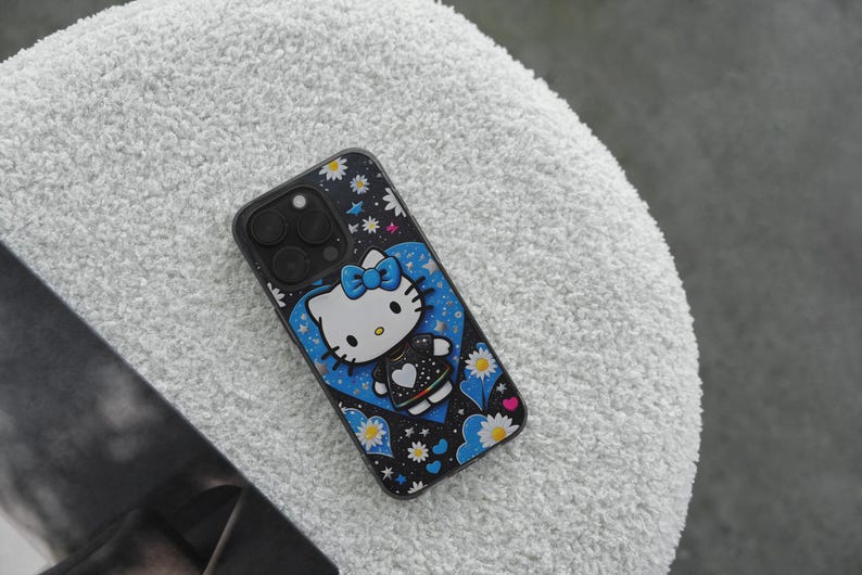 May include: Black phone case with a Hello Kitty design. The case has a black background with white daisies and stars. Hello Kitty is in a blue heart with a blue bow. The case is on a white, textured surface.