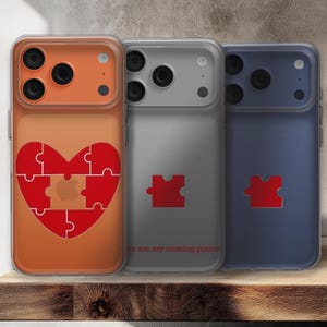 Pode incluir: Três capas de telefone em laranja, cinza e azul. A capa laranja apresenta um coração de quebra-cabeça vermelho com um logotipo da Apple. A capa cinza tem uma peça de quebra-cabeça vermelha e o texto "You are my missing piece". A capa azul tem uma peça de quebra-cabeça vermelha.