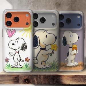 Op de afbeelding: Drie telefoonhoesjes met Snoopy-illustraties. De eerste hoes toont Snoopy met een hart en zon, de tweede met een bloem en de derde met een pot. Elke hoes heeft een transparante rand en een gekleurde bovenkant.