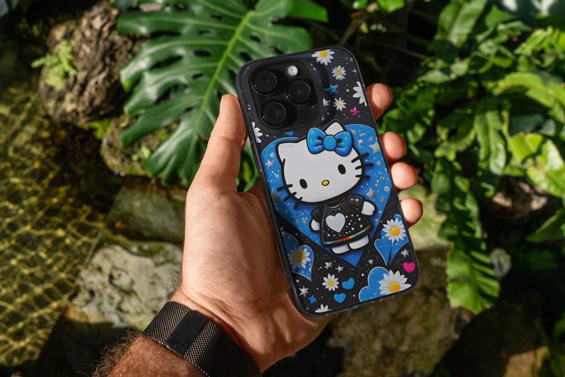 May include: Black phone case with a Hello Kitty design. The case has a black background with white daisies and stars. Hello Kitty is wearing a black dress with a white heart and a blue bow. The case is held in a hand.