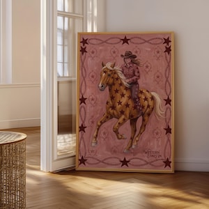 May include: A framed rectangular wall art print featuring a brown horse with dark brown star patterns and a rider wearing a cowboy hat. The background is dusty pink with a dark red rope border and star accents. The text Western Stars 24 is visible.