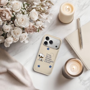 May include: A beige iPhone case featuring Snoopy holding a cross, blue stars, and the text "I can do all things through Christ." The case is surrounded by flowers, candles, and a pen.
