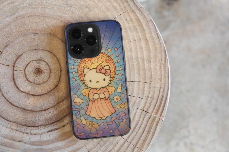 May include: A black phone case with a stained-glass effect, depicting Hello Kitty as an angel. The design includes a halo, wings, and a pink dress against a blue backdrop. The phone case is on a wooden surface.