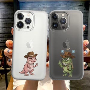 May include: Two phone cases with cartoon illustrations. One case is clear with a pink dinosaur wearing a cowboy hat. The other case is gray with a green frog wearing a cowboy hat and giving the middle finger.