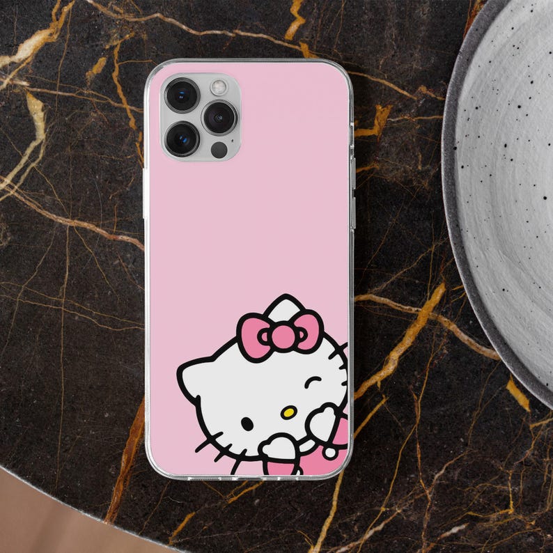 Puede incluir: Funda de tel&eacute;fono rosa con dise&ntilde;o de Hello Kitty. La funda tiene un borde transparente y un fondo rosa claro. El gr&aacute;fico de Hello Kitty incluye un lazo rosa, una cara blanca y contornos negros. La funda est&aacute; sobre una superficie de m&aacute;rmol oscuro.