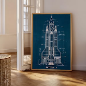 May include: A framed blueprint-style print of the Saturn V rocket against a dark blue background. The white detailed illustration shows the rocket's components and is labeled "SATURN V". The print is in a wooden frame and displayed indoors.