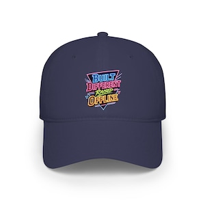 May include: A navy blue baseball cap featuring a neon-style graphic print on the front. The design includes the text BUILT DIFFERENT RAISED OFFLINE in blue, pink, green, and yellow, surrounded by geometric lines in a retro aesthetic style.