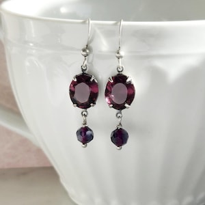 May include: A pair of dangle earrings with silver-tone hooks. Each earring features a large, oval, deep purple gemstone, with a smaller, round, dark purple bead hanging below. The earrings are displayed in front of a white teacup.