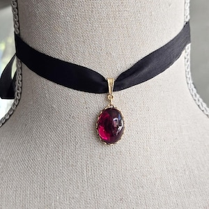 May include: A black satin choker necklace with a gold pendant featuring a large, oval, deep red gemstone. The pendant is suspended from a delicate gold bail. The necklace is displayed on a mannequin.