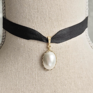 May include: A black ribbon choker necklace featuring a central oval-shaped white pearl pendant set in a gold-toned metal frame. The pendant hangs from a gold-toned bail attached to the smooth black fabric band, creating a simple and classic neck accessory.