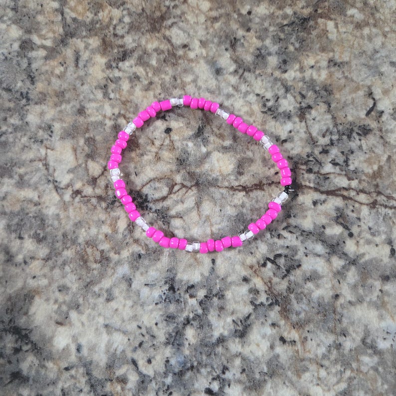 May include: A beaded bracelet featuring bright pink and silver beads. The bracelet is circular and appears to be made of small, evenly spaced beads. The beads are strung together to form a flexible band.