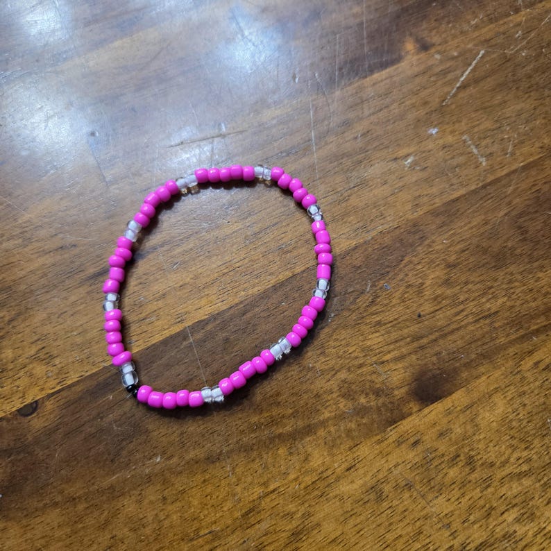 May include: A beaded bracelet featuring vibrant pink beads interspersed with clear and silver beads. The bracelet is circular and rests on a wooden surface, showcasing its design and color contrast.