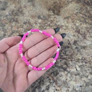 May include: A beaded bracelet featuring bright pink and clear beads. The bracelet is made of small, round beads strung together to form a circular shape. The bracelet is shown on a hand.