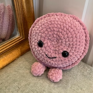 Puede incluir: Un peluche de pulpo rosa tejido a mano con ojos de seguridad negros y una boca sonriente. El peluche tiene un cuerpo redondo y cuatro pequeños tentáculos. El pulpo está sentado sobre una superficie texturizada.