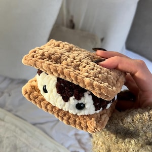 May include: A crocheted ice cream sandwich with a light brown, textured exterior and a white and dark brown filling. The sandwich has black bead accents and is held in a hand.