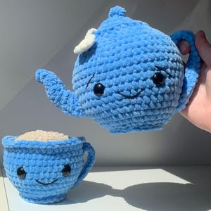 Könnte beinhalten: Ein gehäkeltes blaues Teekannen- und Tassenset mit lächelnden Gesichtern und schwarzen Knopfaugen. Die Teetasse ist mit einer beigen Substanz gefüllt, die möglicherweise Tee oder Kaffee darstellt. Die Teekanne hat einen weißen Akzent oben.