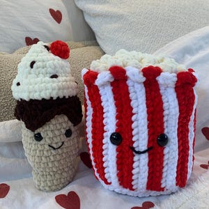 May include: Crocheted cupcake and popcorn bucket. The cupcake has a white top with chocolate chips and a red cherry. The popcorn bucket is red and white striped with a smiling face and filled with white popcorn.