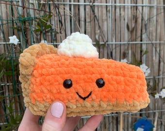 May include: A crocheted slice of pie with a smiling face and a dollop of white cream. The pie has an orange filling and a tan crust. The pie is a handmade item.