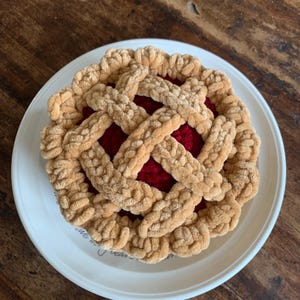 May include: A crocheted pie on a white plate. The pie has a lattice crust in a light brown colour, covering a red filling. The plate has a simple design and sits on a wooden surface.