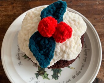 May include: A crocheted Christmas pudding with white, red, and teal accents, resting on a decorative plate. The pudding features a white top, a brown base, and is adorned with red pom-poms and teal leaves. The plate has text that reads "The holly and..."
