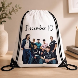 Puede incluir: Una mochila de cordón blanca con un estampado gráfico negro de un grupo de seis jóvenes posando juntos. Sobre el grupo, el texto "December 10" está impreso en una fuente cursiva negra. La bolsa tiene cordones negros y detalles en las esquinas.