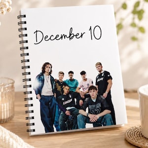Könnte beinhalten: Ein weißes Notizbuch mit Spiralbindung, auf dessen Vorderseite oben der Text December 10 in schwarzer Schreibschrift gedruckt ist, darunter befindet sich ein Foto einer Gruppe von sechs jungen Menschen in Freizeitkleidung.