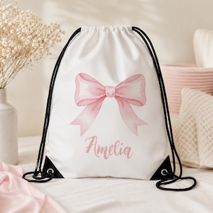 May include: A white drawstring backpack featuring a large, watercolour-style pink bow printed in the centre. Below the bow, the name Amelia is written in a cursive pink script. The bag has black cord straps and black reinforced corner patches.