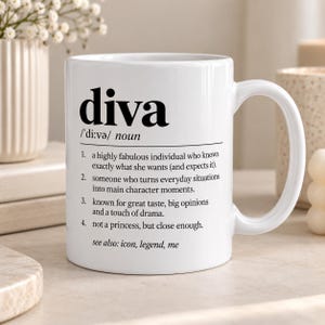 May include: A white ceramic mug featuring the dictionary definition of the word diva in black text. The print includes the phonetic spelling, the noun classification, and four numbered definitions describing a fabulous individual, followed by see also: icon, legend, me.