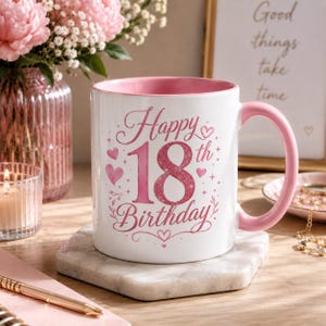 Könnte beinhalten: Ein weißer Keramikbecher mit rosafarbenem Innenraum und Henkel, der auf der Vorderseite den rosafarbenen Glitzertext Happy 18th Birthday zeigt, umgeben von kleinen rosa Herzen und dekorativen Verzierungen.