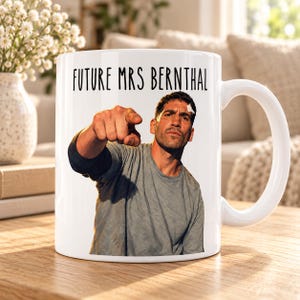 Pode incluir: Uma caneca de cerâmica branca com a imagem impressa de um homem a apontar para a frente e com um penso na testa. Por cima da imagem, o texto FUTURE MRS BERNTHAL está escrito numa fonte preta sem serifa. A caneca tem uma pega padrão.