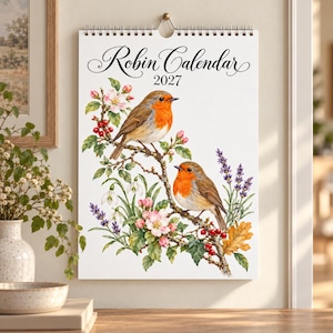 May include: A 2027 wall calendar titled Robin Calendar featuring a white background with two illustrated robins perched on flowering branches. The artwork includes pink blossoms, red berries, lavender sprigs, and green leaves.