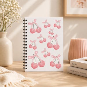 Pode incluir: Um caderno de espiral branco com um padrão repetitivo de cerejas cor-de-rosa em estilo aguarela. Cada par de cerejas está ligado por um delicado laço de fita rosa. A capa apresenta um design limpo e simples com várias ilustrações.