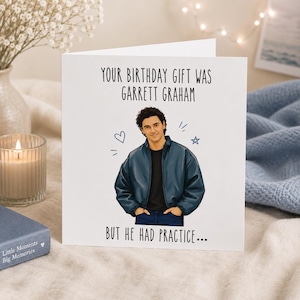 May include: A white greeting card featuring a cartoon illustration of a person with curly dark hair wearing a dark blue jacket over a black shirt. The text reads: YOUR BIRTHDAY GIFT WAS GARRETT GRAHAM BUT HE HAD PRACTICE... with a small heart and star.