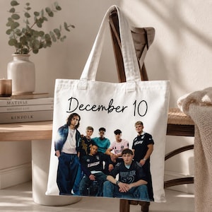 May include: A white canvas tote bag featuring the black text "December 10" at the top and a photographic print of a group of six young people posing together. The individuals are wearing casual clothing including t-shirts, jackets, and jeans.