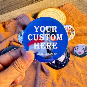 Op de afbeelding: Een verzameling aangepaste buttons met diverse ontwerpen. De centrale button is blauw met de tekst "YOUR CUSTOM HERE" en een pinguïn-afbeelding. Andere buttons tonen verschillende illustraties. De buttons liggen op een oranje stoffen achtergrond.