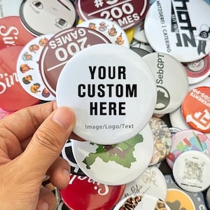 May include: A collection of various round buttons, some with text and graphics. A large white button in the center displays "YOUR CUSTOM HERE" in black, with "Image/Logo/Text" below. Other buttons feature text like "200 GAMES" and "METZGEREI | CATERING".