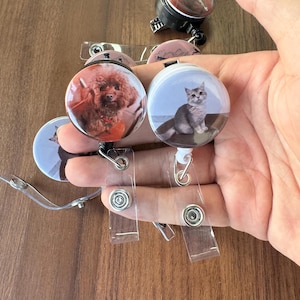 May include: A collection of retractable badge holders featuring pet portraits. One shows a brown poodle, another a gray and white cat. Each has a clear plastic strap and a metal clip for attaching to clothing.