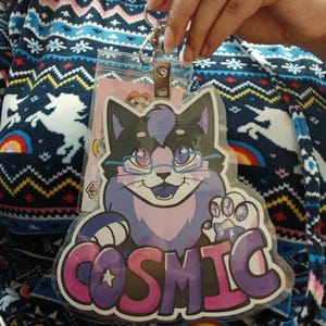 May include: A clear plastic badge holder with a cartoon cat design. The cat is black, purple, and blue, wearing glasses, and holding a paw with stars. The word "COSMIC" is written in pink and purple letters.