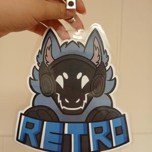 May include: A badge featuring a stylized blue and black wolf-like character with the word "RETRO" in blue block letters. The badge has a clear plastic covering and a metal clip.
