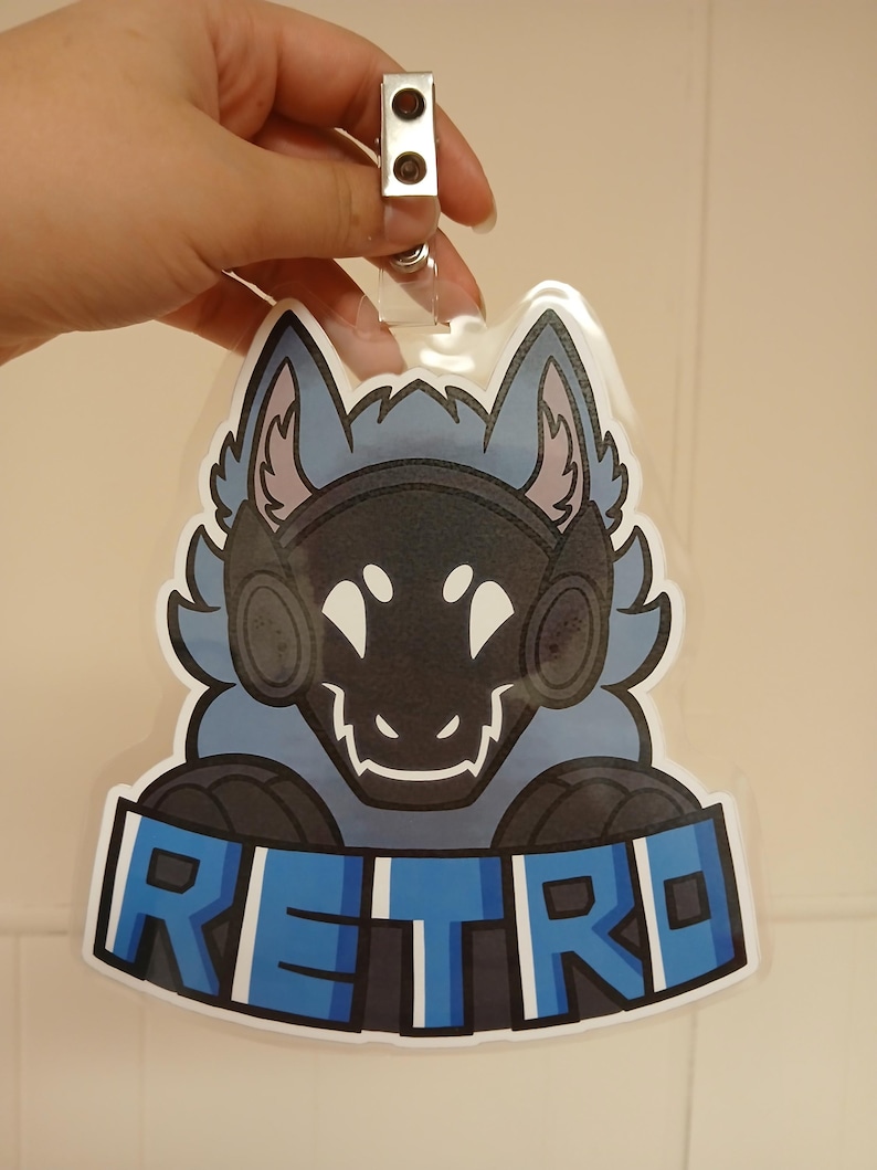 May include: A badge featuring a stylized blue and black wolf-like character with the word "RETRO" in blue block letters. The badge has a clear plastic covering and a metal clip.