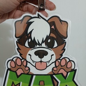 May include: A clear plastic badge featuring a cartoon dog with brown and white fur, large black eyes, and a pink tongue. The name "MAX" is displayed in green block letters at the bottom. A metal clip is attached at the top.