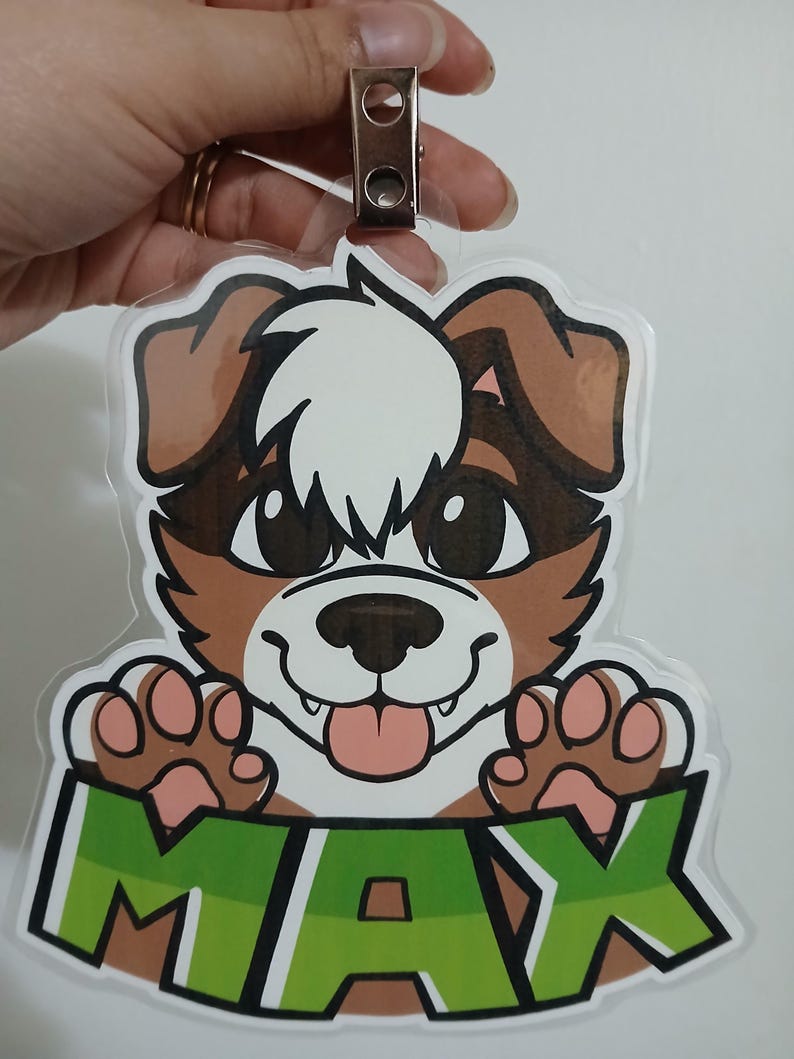 May include: A clear plastic badge featuring a cartoon dog with brown and white fur, large black eyes, and a pink tongue. The name "MAX" is displayed in green block letters at the bottom. A metal clip is attached at the top.