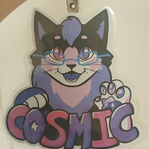 May include: A clear plastic badge featuring a cartoon cat with black, purple, and pink fur, wearing glasses. The badge reads "COSMIC" in a gradient of pink and purple. A metal clip and lanyard are attached at the top.