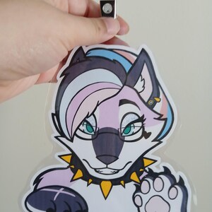 May include: A clear plastic badge featuring a cartoon wolf with a trans pride color scheme. The wolf has a black nose, teal eyes, and a spiked collar. The badge is held by a clear plastic clip.