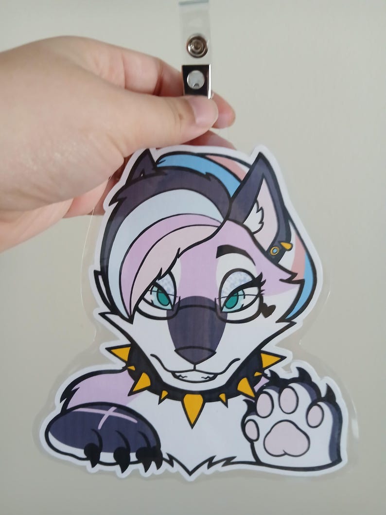 May include: A clear plastic badge featuring a cartoon wolf with a trans pride color scheme. The wolf has a black nose, teal eyes, and a spiked collar. The badge is held by a clear plastic clip.