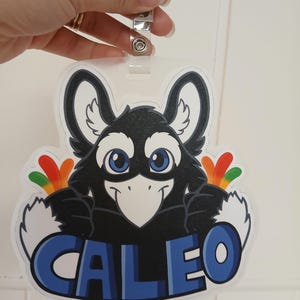 May include: A clear plastic name tag with a cartoon bird design. The bird is black with white accents, blue eyes, and colorful feathers. The name "CALEO" is displayed in blue letters.