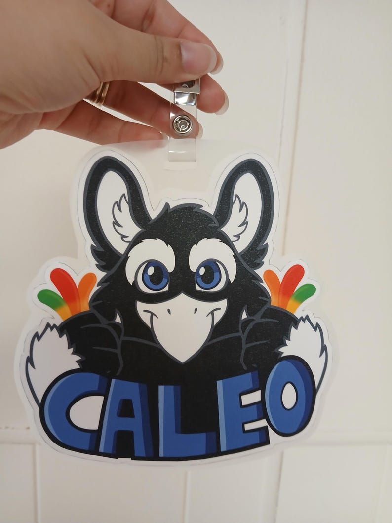 May include: A clear plastic name tag with a cartoon bird design. The bird is black with white accents, blue eyes, and colorful feathers. The name "CALEO" is displayed in blue letters.