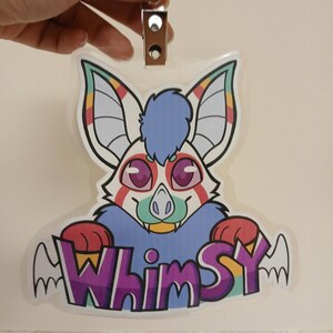 May include: A clear, plastic badge featuring a cartoon bat with large ears and a blue tuft of fur. The bat has pink eyes, a green nose, and a smiling mouth. The word "Whimsy" is written in purple and yellow letters with wing accents.